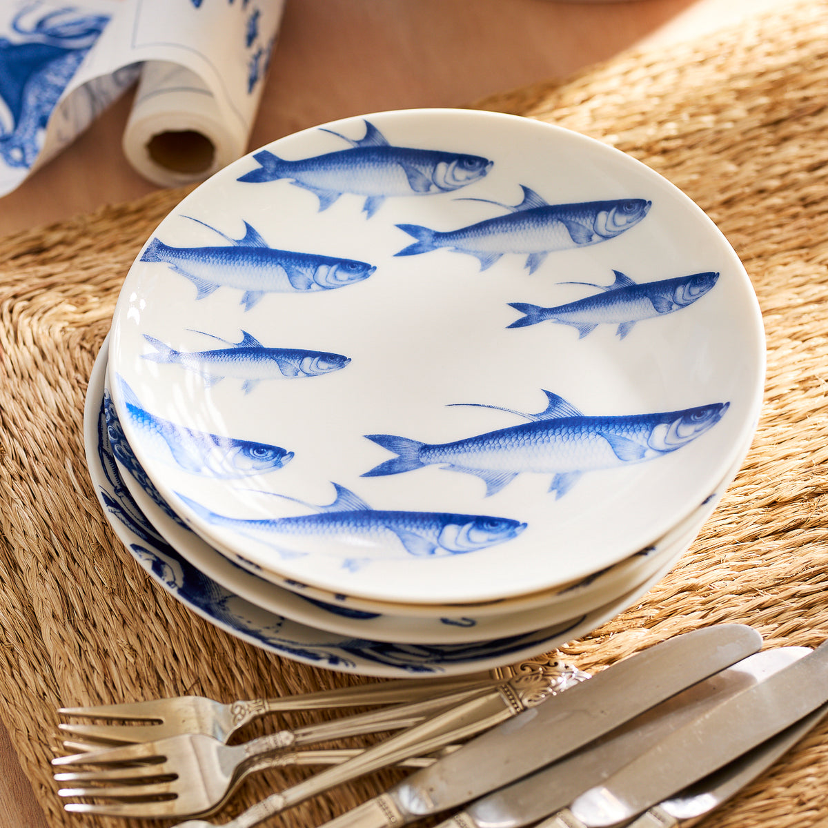 Stack of Caskata Lake House Bundle plates decorated with a school of fish designs, set on a woven placemat beside silver forks and knives, perfect for your lake house.
