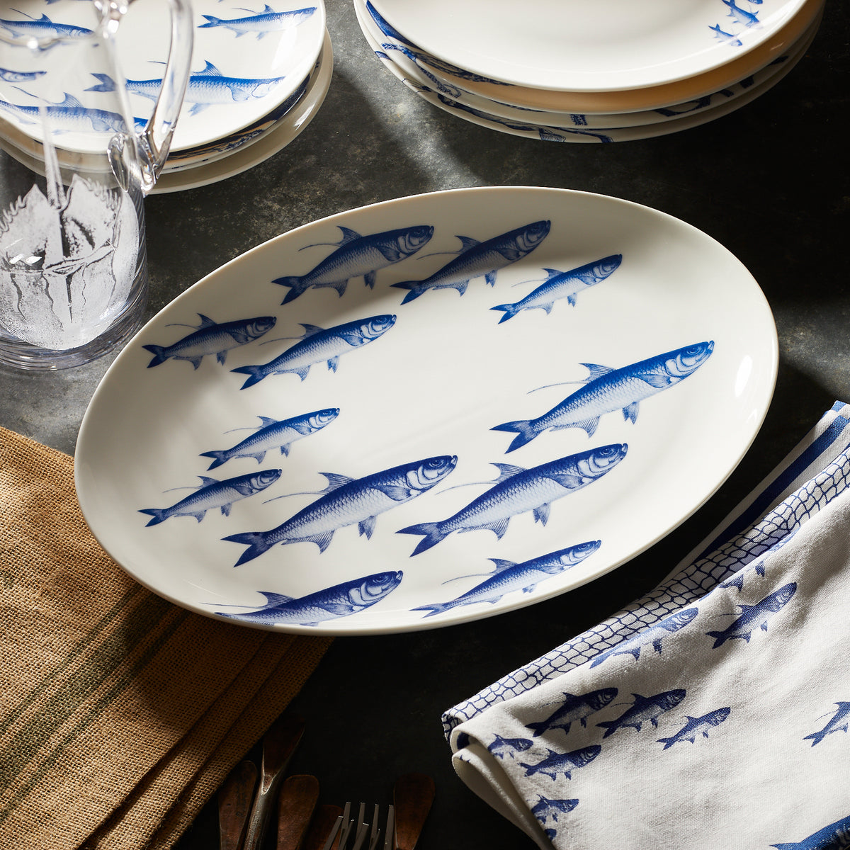 A white oval dish featuring a blue fish pattern is displayed on a dark surface, accompanied by a folded napkin with a similar fish design and other stacked plates in the background. This ensemble is part of the Lake House Bundle by Caskata.