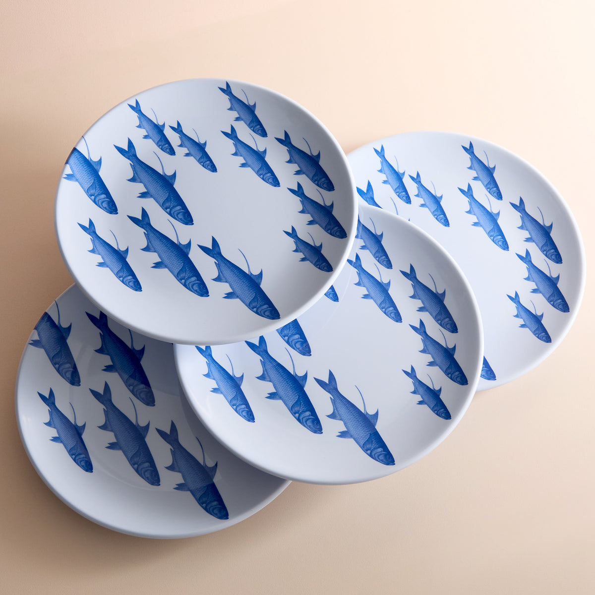 A set of four &quot;School of Fish Melamine Dinner Plates&quot; by Caskata, featuring blue fish patterns, is arranged diagonally on a beige surface. These plates add seaside charm to your table setting.