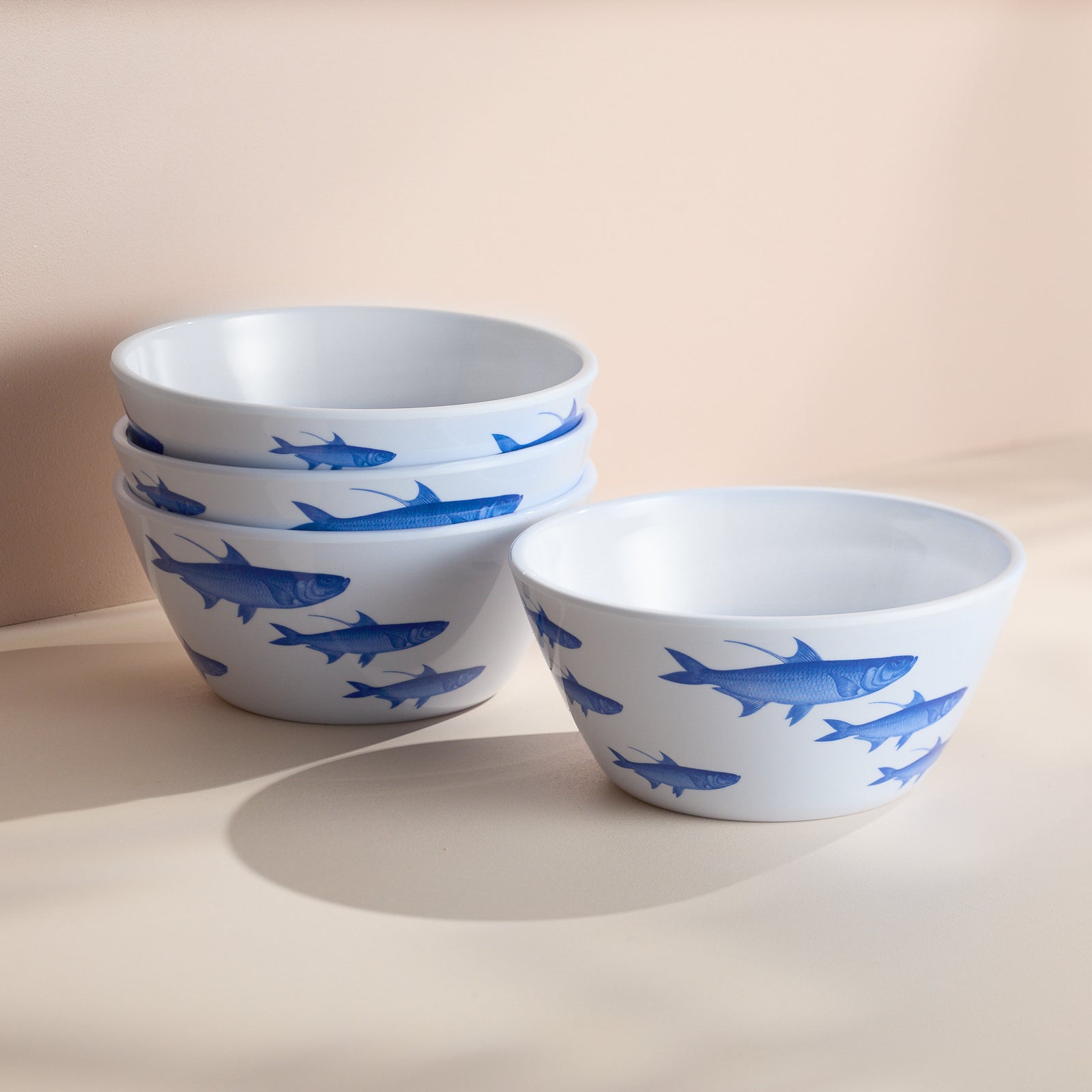 Four elegant Caskata School of Fish Melamine Cereal Bowls with blue fish designs are stacked on a beige surface, offering both style and function for your outdoor collection.