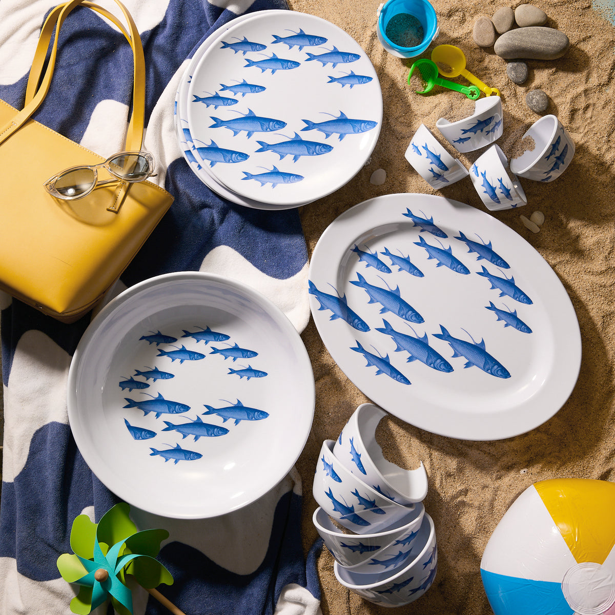 A vibrant beach scene features Caskata’s &quot;School of Fish&quot; Melamine Oval Rimmed Platter, a bright yellow bag with sunglasses, a striped towel, rocks, colorful toys, and a playful beach ball on the sand.