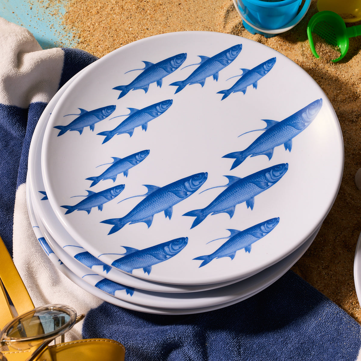 Caskata&#39;s School of Fish Melamine Dinner Plates, Set of 4, rests on sandy ground alongside a dark towel, sunglasses, and beach toys—perfectly embodying an outdoor collection made from BPA-free melamine.