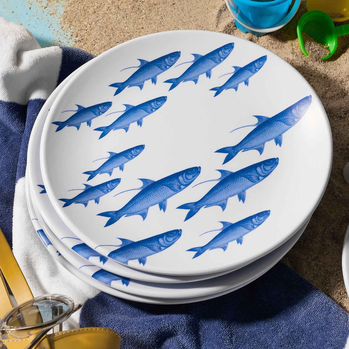 A stack of Caskata&#39;s School of Fish Melamine Dinner Plates, Set of 4, rests on sand alongside a towel, sunglasses, and beach toys.