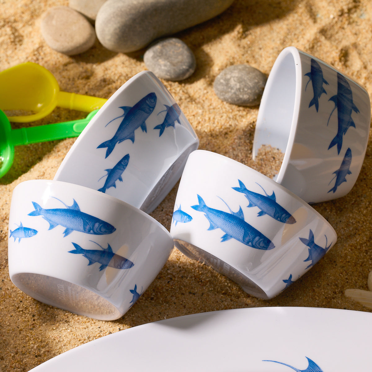 A set of four Caskata School of Fish Melamine Snack Bowls sits on sandy ground among stones and colorful toy shovels.