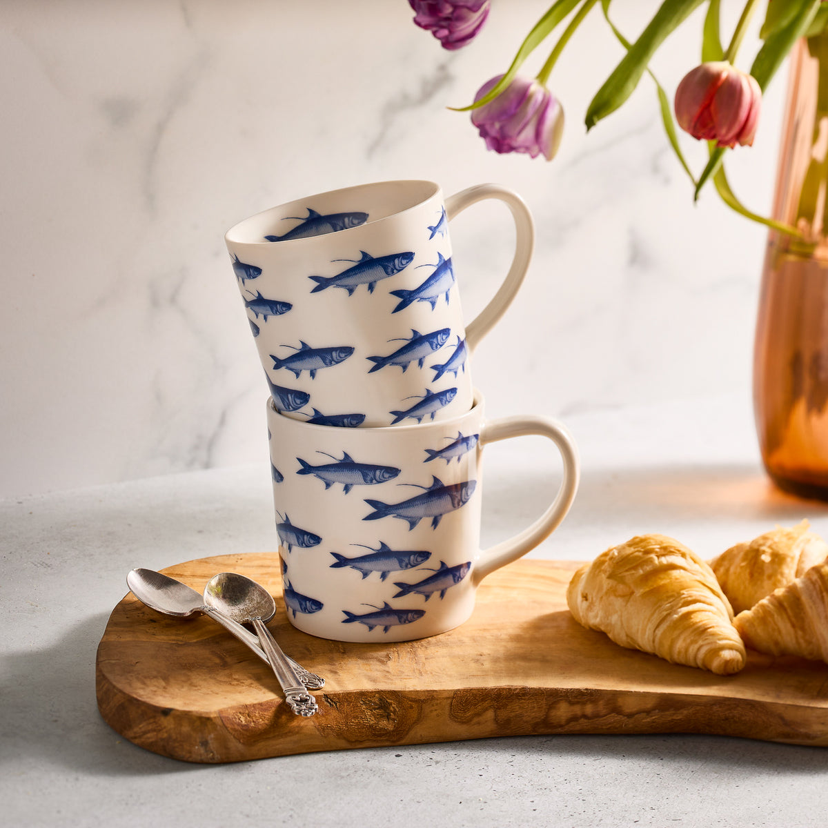 Two Caskata Artisanal Home School of Fish Mugs, crafted from high-fired porcelain and adorned with blue fish patterns, are stacked on a wooden board with croissants, spoons, and a vase of flowers in the background.