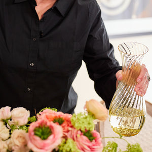 A person in a black shirt is pouring liquid from a Quinn Citrine Carafe by Caskata Artisanal Home into a vase near assorted colorful flowers.