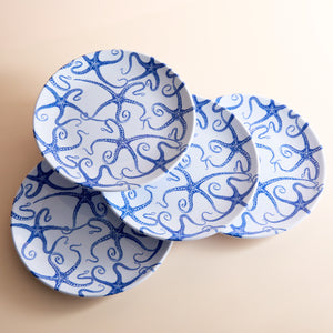 On a light beige surface, four Caskata Starfish Melamine Dinner Plates feature a charming blue starfish pattern. Crafted from BPA-free melamine, these plates blend durability with seaside elegance.