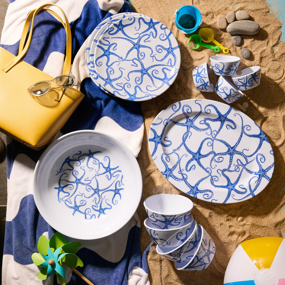 A beach scene with the Caskata Starfish Melamine Oval Rimmed Platter showcases blue and white starfish patterns, paired with a yellow bag, sunglasses, striped towel, beach ball, and small toys on the sand—a perfect setup for outdoor entertaining.