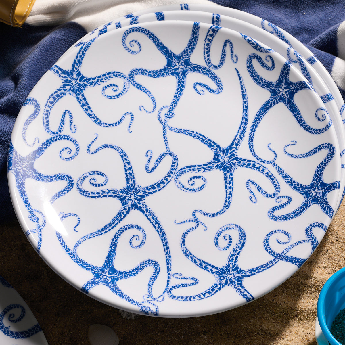 The Starfish Melamine Dinner Plates by Caskata feature intricate blue octopus designs on a round white base, ideal for outdoor dining, with hints of dark blue fabric showing through sand. Comes in a set of 4.