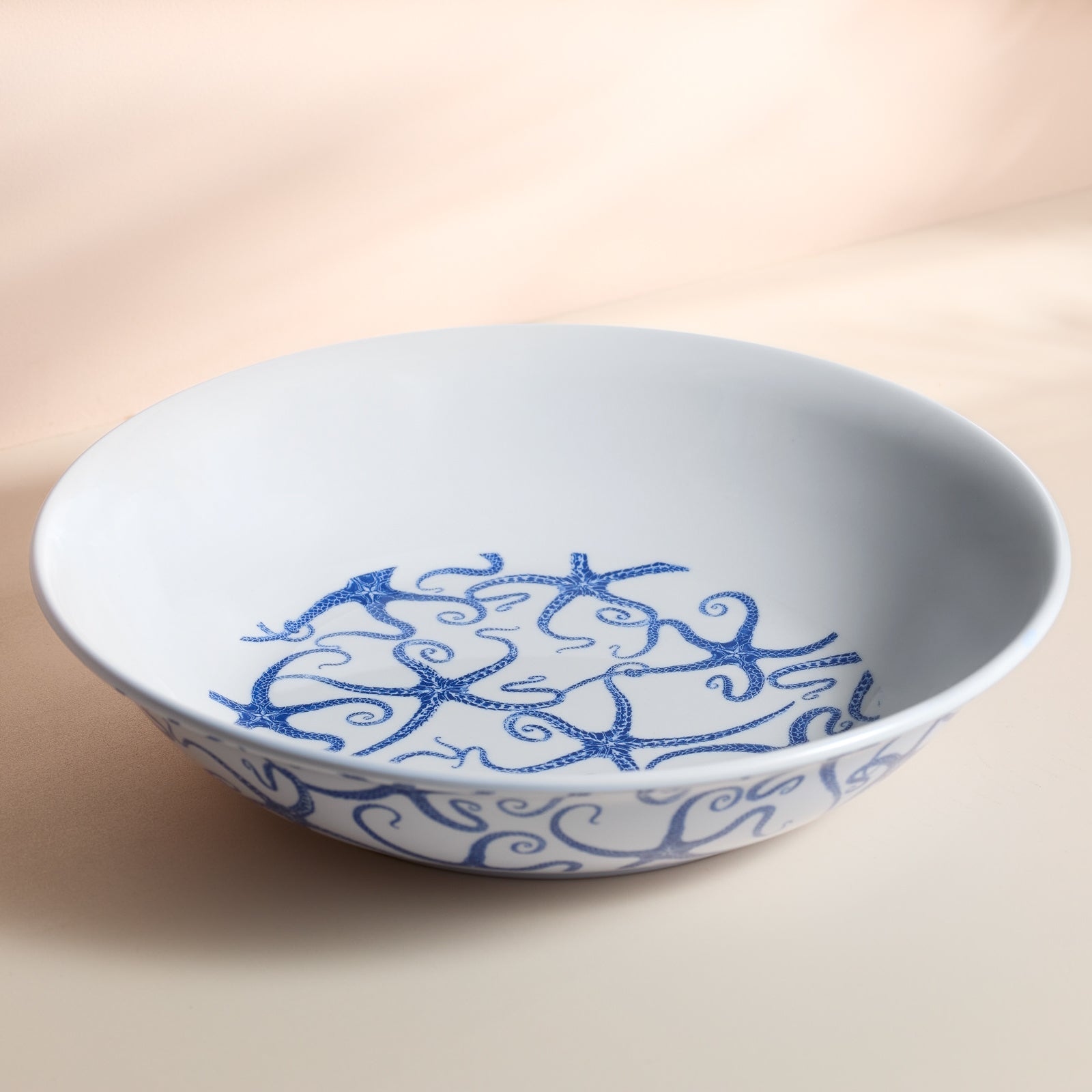 The Caskata Starfish Melamine Serving Bowl, BPA-free and adorned with blue starfish and swirl patterns inside and along the rim, lies on a light beige surface—ideal for outdoor entertaining.