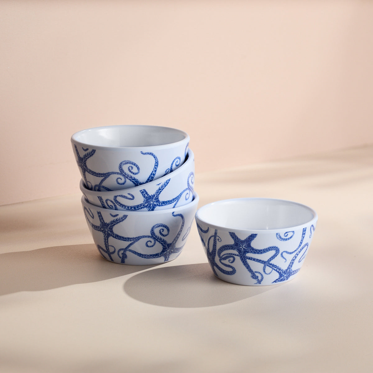 A stack of four Caskata Starfish Melamine Snack Bowls featuring a starfish design sits on a beige surface, with one separated on the right. These BPA-free bowls combine unbreakable style and practicality for any setting.