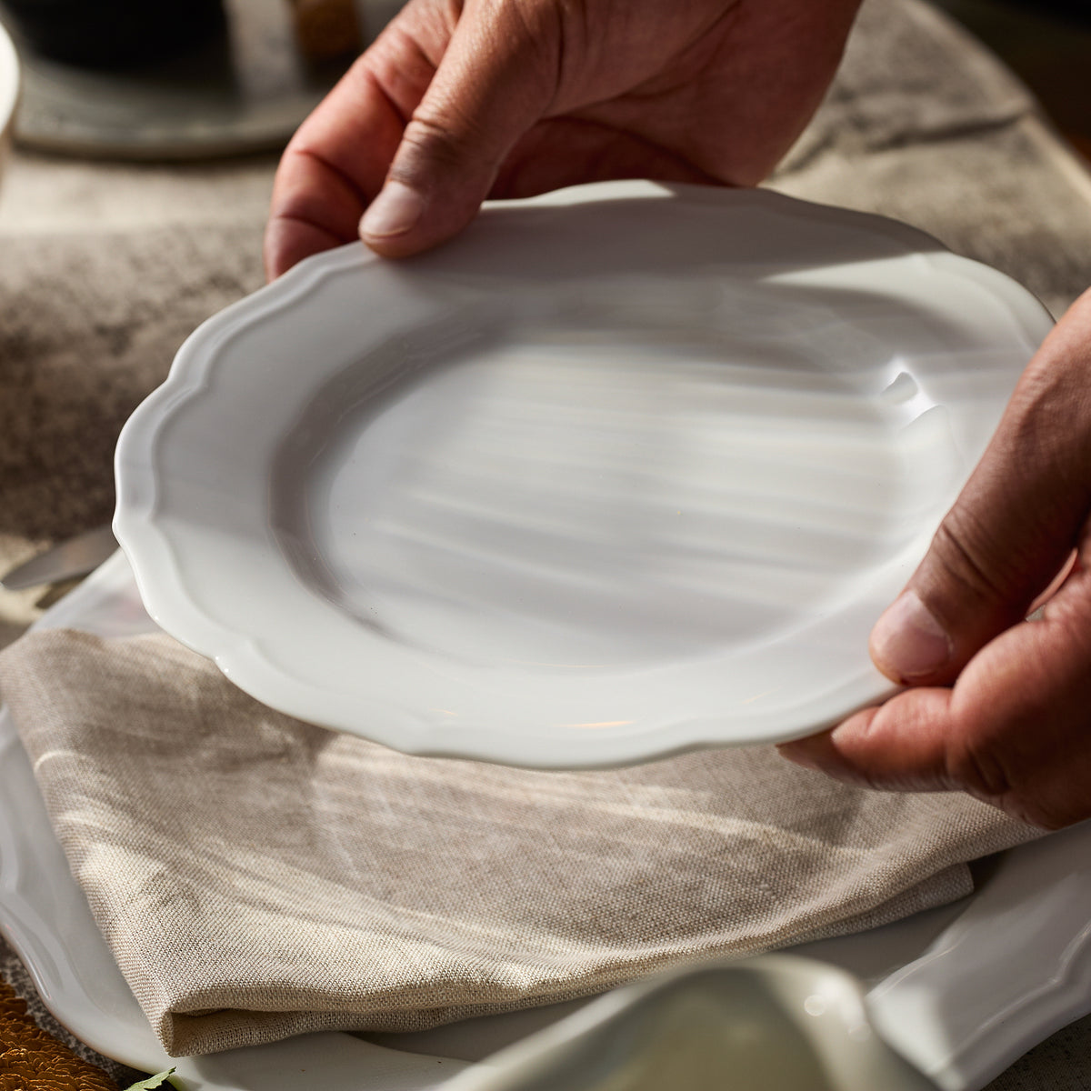 Hands carefully placing a Caskata Stella Scalloped White Salad Plate on a beige cloth, evoking the charm of vintage dinnerware.