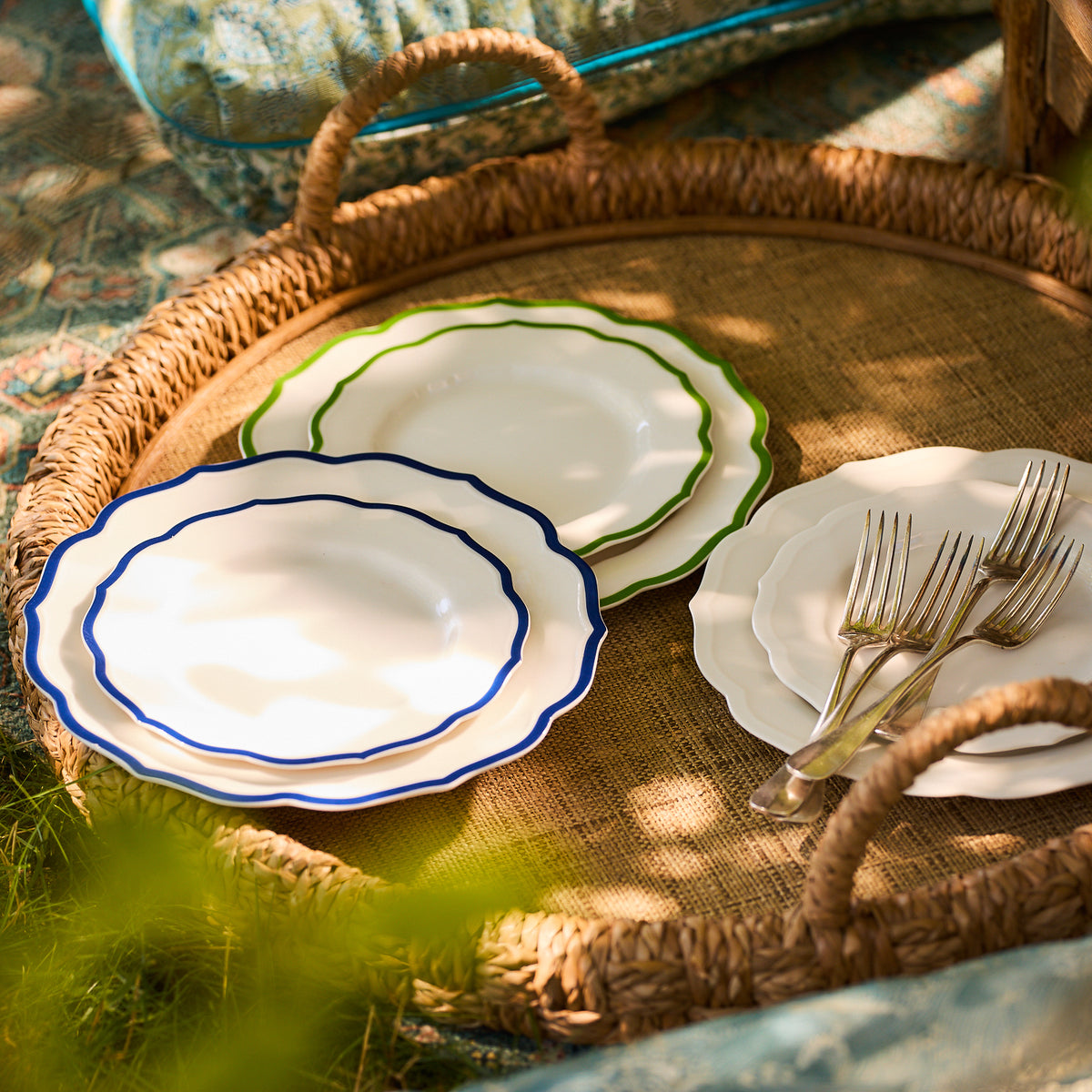 A wicker tray from the Caskata collection holds three stacked Stella Scalloped Verde Salad Plates, their neutral palette showcasing green, blue, and white borders. Four forks rest beside the plates, exuding an heirloom feel in this charming outdoor setting.