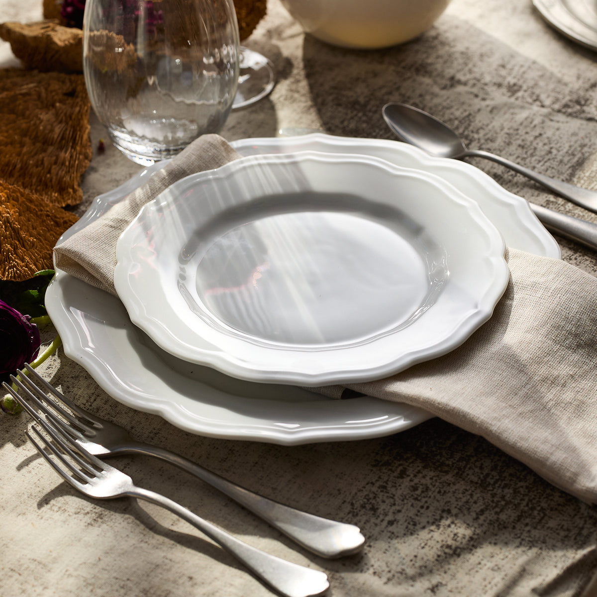 An elegant table setting includes a vintage dinner plate, the Stella Scalloped White Dinner Plate by Caskata, a folded beige napkin, silverware, and a glass on a textured surface.