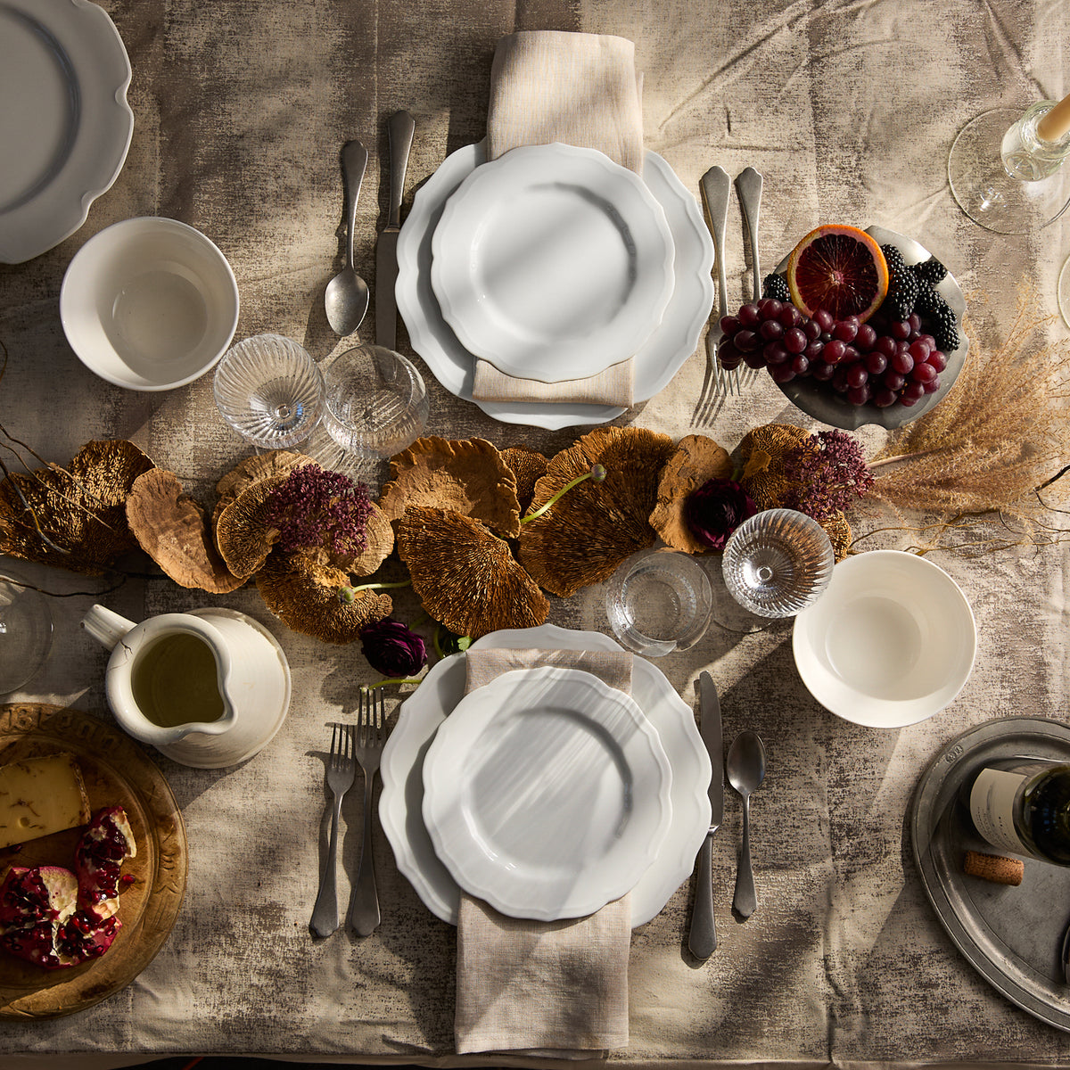 A rustic table setting features Caskata&#39;s Stella Scalloped White Dinner Plate, beige napkins, and assorted glasses, surrounded by dried leaves and flowers. A vintage touch is added with a platter of pomegranates and a dish of grapes, completing the charming display.