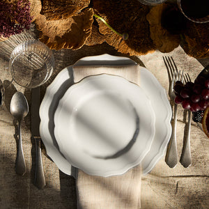 A table setting with a Stella Scalloped White Dinner Plate by Caskata, a beige napkin, silver cutlery, a glass, and grapes on a rustic tablecloth. Sunlight casts shadows across the scene.