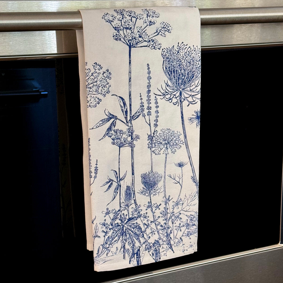 A white cotton kitchen towel adorned with blue botanical illustrations from the Summer Blues Kitchen Towels set by Caskata is hanging on an oven handle.