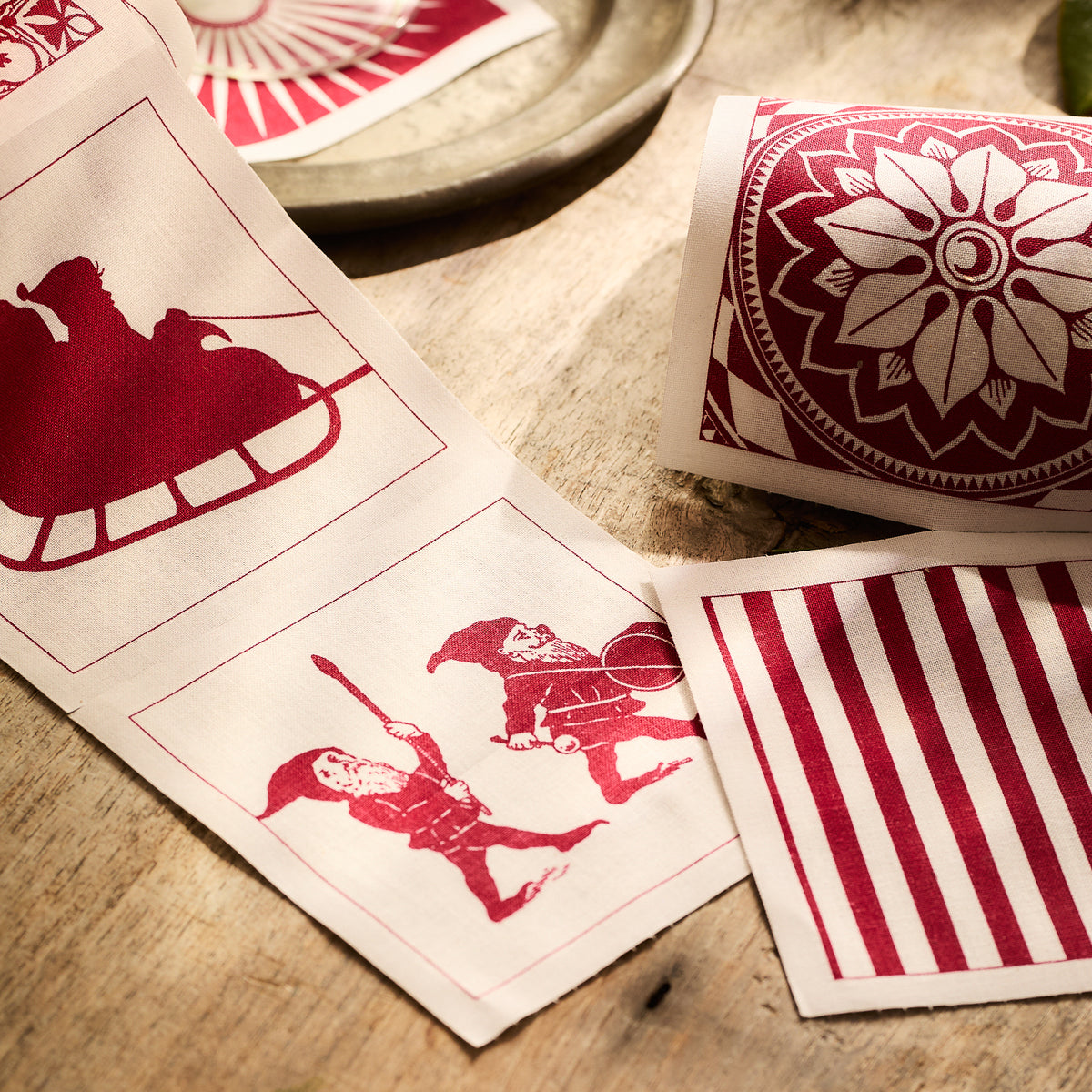 The Holiday Fun Cocktail Napkin Rolls, Set of 2 by MY DRAP feature a red and white textile pattern with Santa&#39;s sleigh, floral motifs, two gnomes, and stripes on a wooden surface. These reusable napkins are the perfect festive accessory for cocktail occasions.