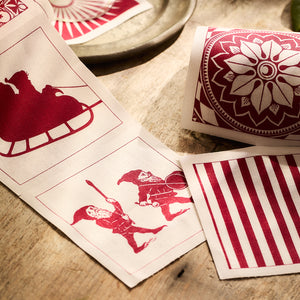 The Holiday Fun Cocktail Napkin Rolls, Set of 2 by MY DRAP feature a red and white textile pattern with Santa's sleigh, floral motifs, two gnomes, and stripes on a wooden surface. These reusable napkins are the perfect festive accessory for cocktail occasions.