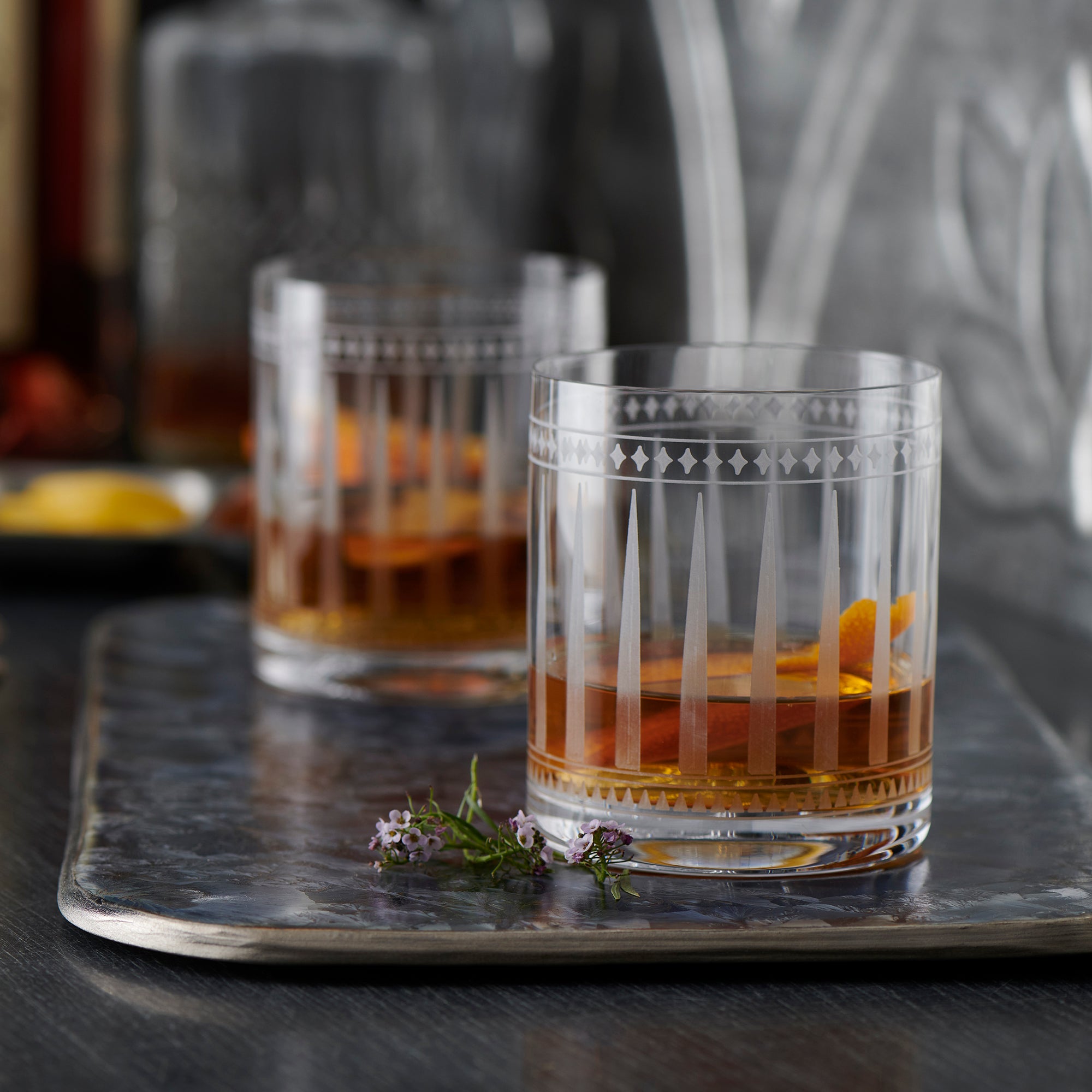 Two clear glass tumblers, crafted using the sand-etching technique, feature geometric etched patterns and a reflective surface. These Marrakech Rocks Glasses from Caskata Artisanal Home evoke an Art Deco crystal design.